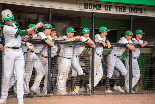 Cary High School players standing against dugout railing looking out towards the field with one player swinging a bat in on-deck circle