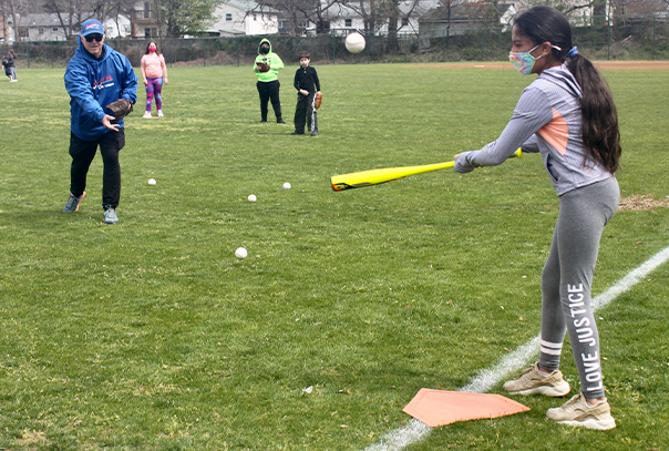 Steve Sternberg in blue sweatshirt underhand tosses baseball to a fourth grade girl swinging a bat with fellow students in the grass field