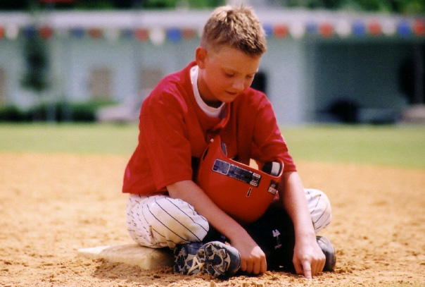 Youth baseball player sitting on base with helmet in his lap and playing in the infield dirt with his hands