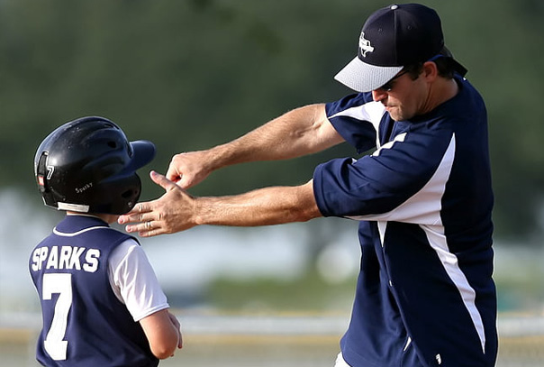 Coach demonstrating a baseball swing to a youth player wearing a helmet a jersey with last name of Sparks on the back