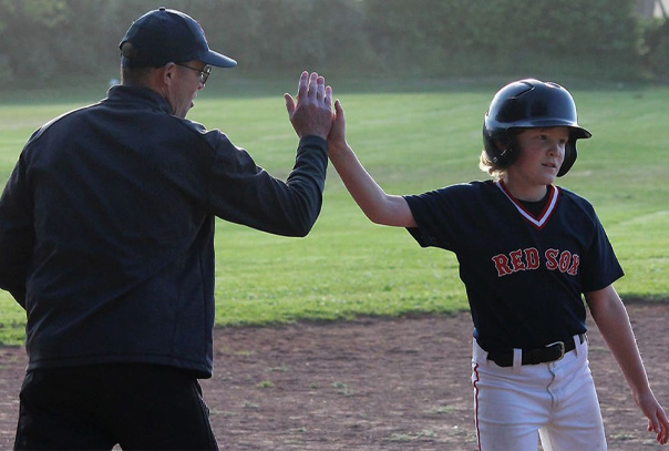 Youth baseball coach in hat and glasses high fives a player wearing a helmet and Red Sox jersey