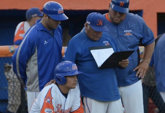 Ron Polk holding clipboard has hand on his player's shoulder who is crouched down with a bat just outside the dugout. Two other coaches are also around them.