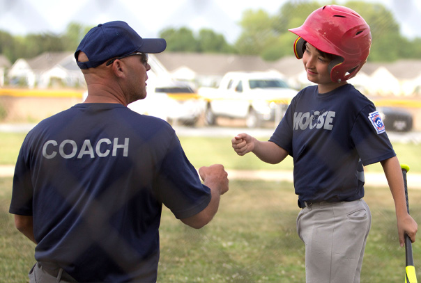 Coach wearing navy blue shirt that says Coach on the back of it giving a fist bump to youth aged player in red helmet walking towards him with a bat in hand.