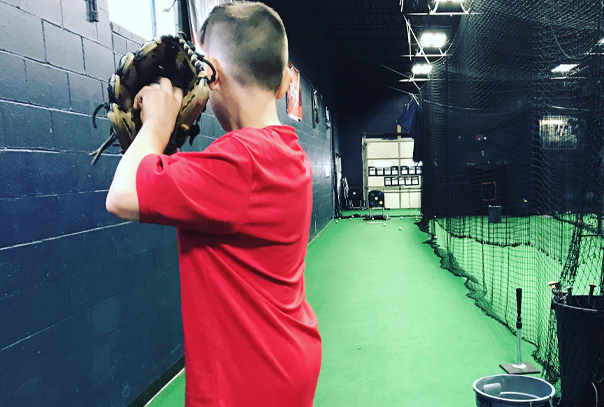 Youth baseball player in red shirt holding glove with ball inside up by his ear as if getting ready to throw a pitch in an indoor facility