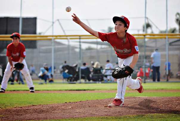 Youth pitcher in red jersey releasing baseball mid-pitch